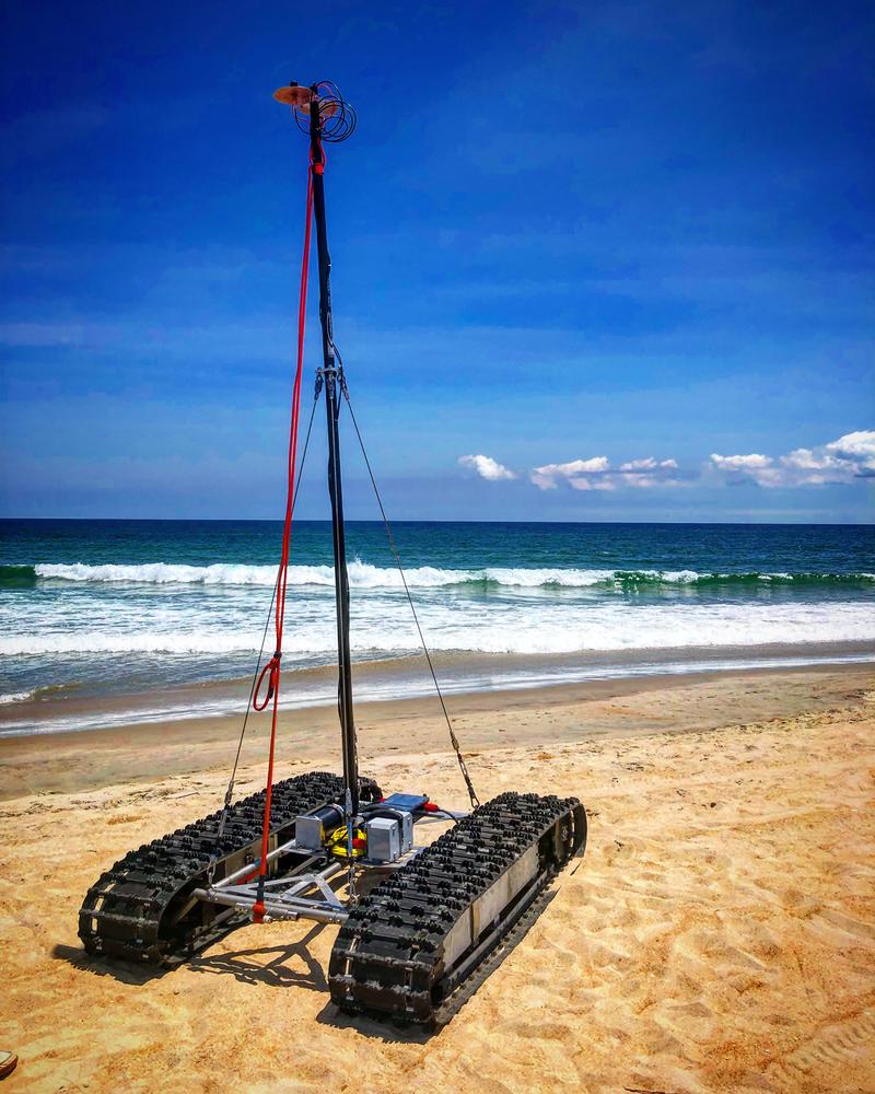 A C2-I Engineering robotic crawler positioned on a coastal beach with its survey mast and antenna deployed